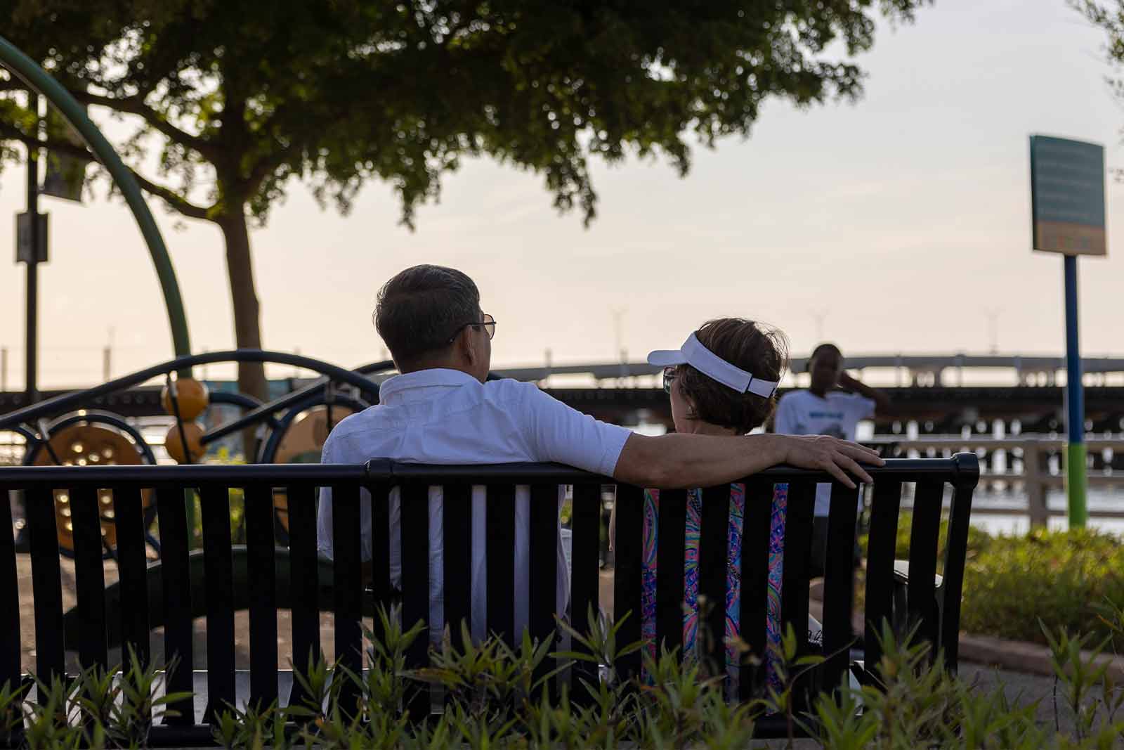 two people sitting on a park bench looking out at the riverwalk and sunset